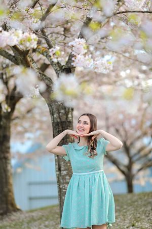 A person in a mint green dress poses playfully under blooming cherry blossom trees in springtime.