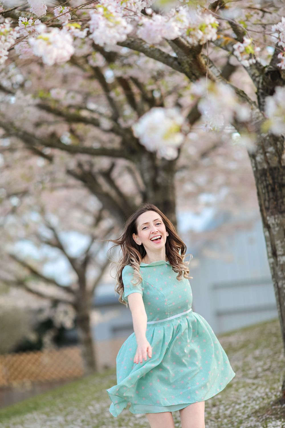 Energetic person in turquoise dress twirls joyfully under blooming cherry blossom trees on a spring day.