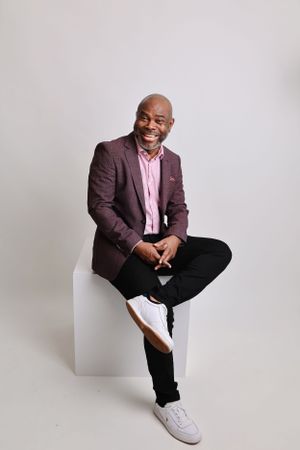 Casual professional portrait in burgundy blazer and white sneakers on white cube.
