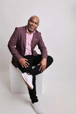Relaxed portrait pose in burgundy blazer and white sneakers against light background.