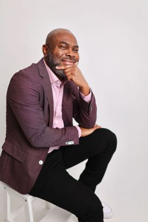 Professional portrait in burgundy blazer and pink shirt sitting on white cube.