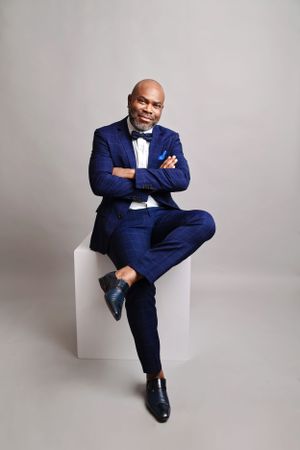 Professional portrait in navy suit and bowtie sitting on white cube against gray background.