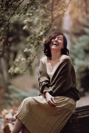 A joyful autumn portrait of someone in a brown sweater and beige skirt sitting on a wooden bench outdoors.