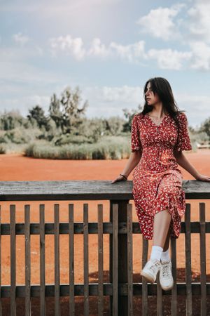 Person in a red floral dress sitting on a wooden railing against a blue sky.