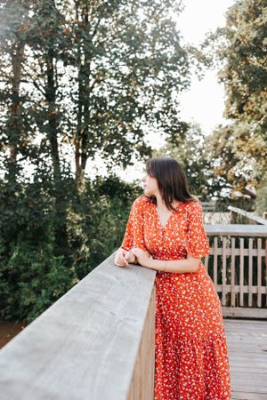 Person in a red floral dress leaning on a wooden deck railing surrounded by trees.