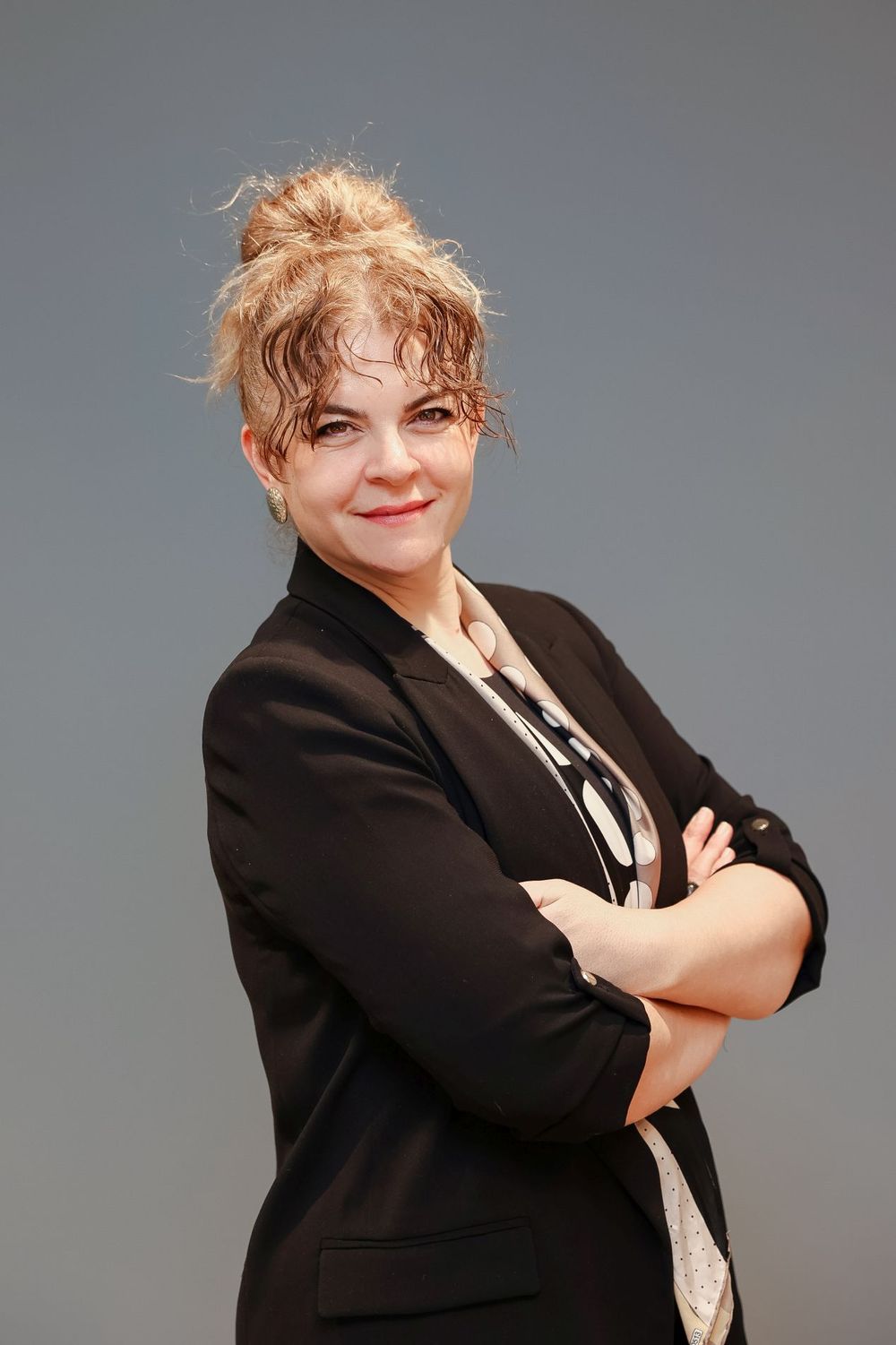 Professional portrait of someone in a black blazer and patterned shirt standing with arms crossed against a gray backdrop.