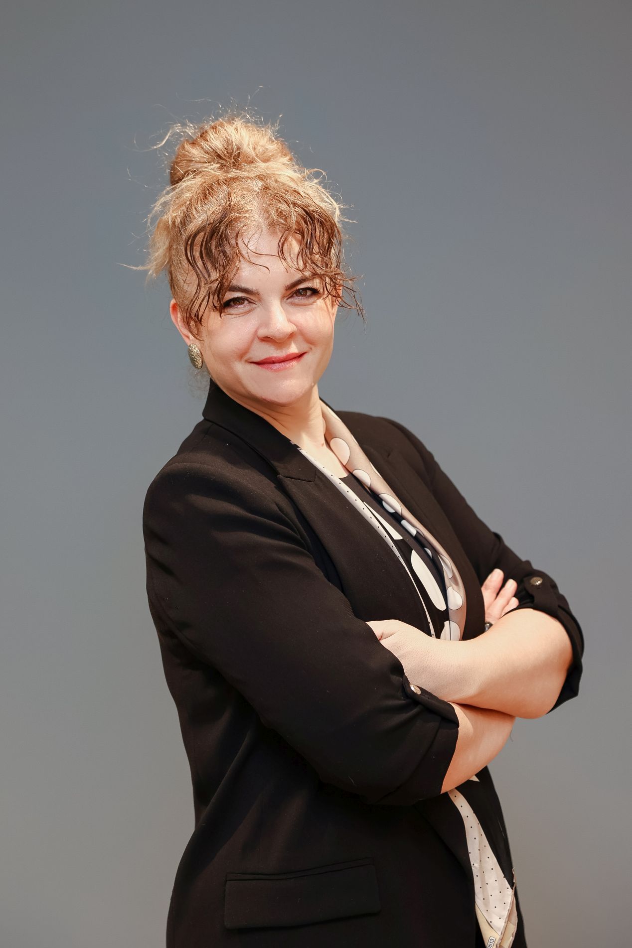 Professional portrait of someone in a black blazer and patterned shirt standing with arms crossed against a gray backdrop.