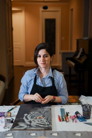 Artist works on charcoal drawing at desk with art supplies and piano visible in background.