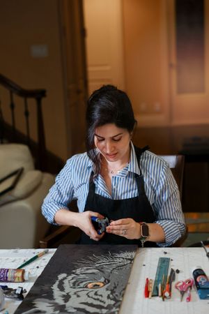 Artist in striped shirt and black apron works on detailed chalk or charcoal tiger drawing at desk with art supplies.
