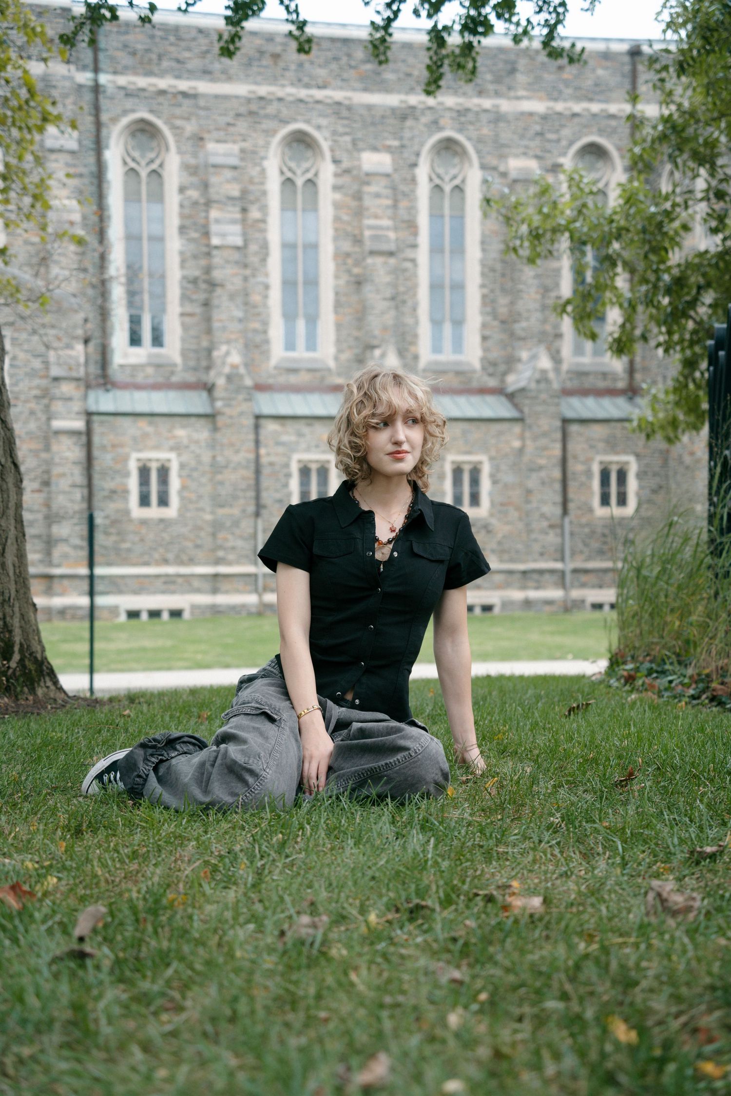 Young woman with curly blonde hair sits cross-legged on grass in front of a stone Gothic university building.