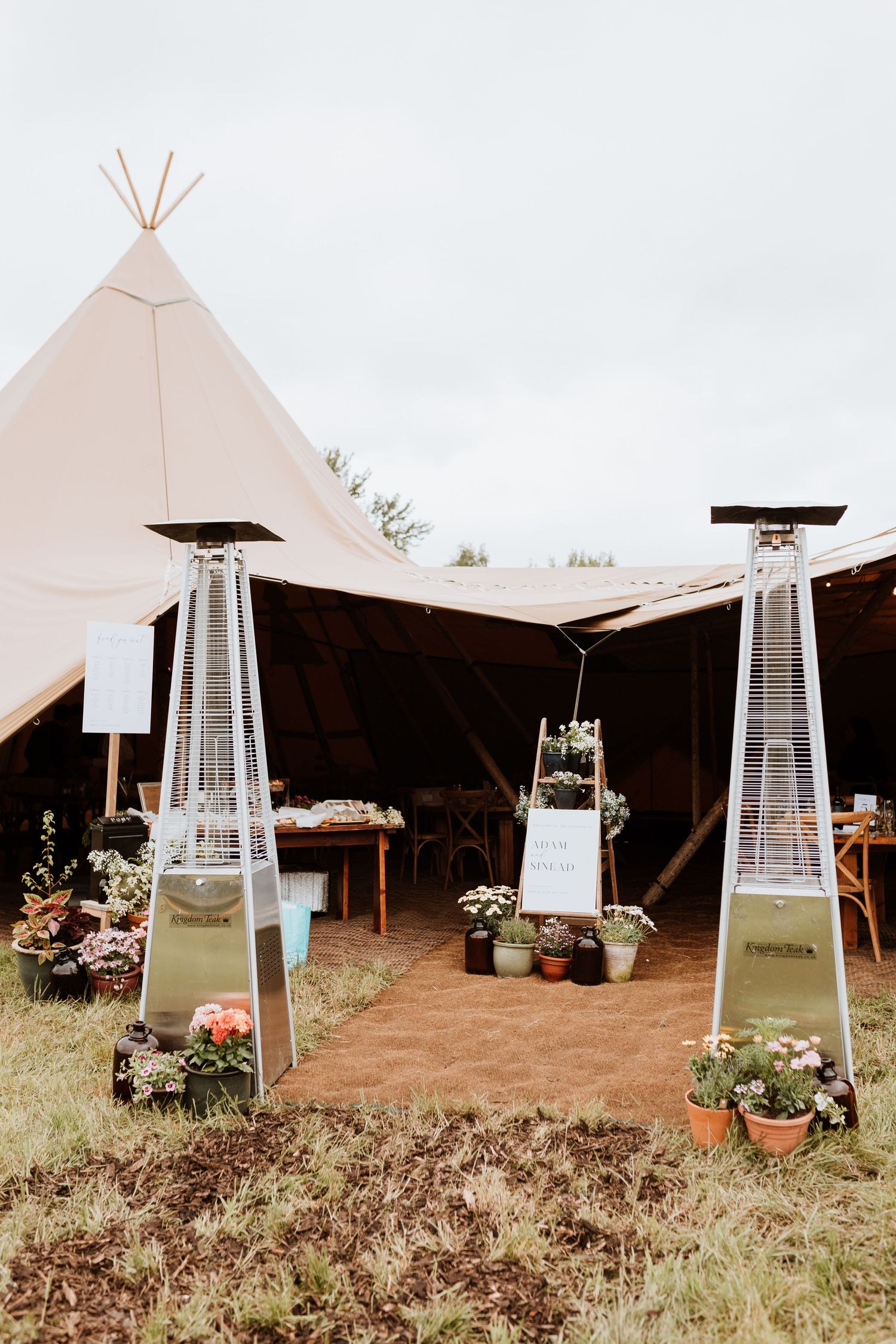 Outdoor event setup with teepee-style tent and tall patio heaters decorated with potted plants and flowers.