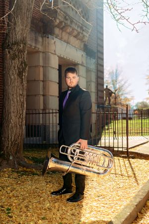 A musician in a formal black suit holds a polished brass euphonium while standing on autumn leaves near a stone building.