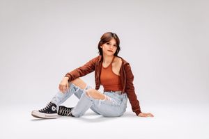 A model in ripped jeans and brown jacket sits casually on white studio floor wearing black and white sneakers.