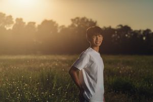 Person wearing a white t-shirt stands in a sunlit meadow at golden hour with trees silhouetted in the background.