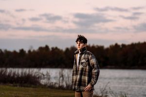 A person in a plaid coat stands near a lake at dusk with fall trees in the background.