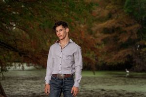 A person stands outdoors in casual attire against a backdrop of autumn foliage in a natural setting.