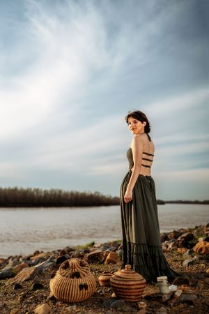 A figure in a flowing dress poses on a rocky shore at sunset with woven baskets beside them.