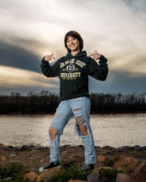 Person wearing a dark university sweatshirt and ripped jeans stands victoriously on rocky terrain at sunset.