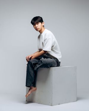 A person wearing a white shirt and jeans sits casually on a white cube against a gray background in a studio setting.