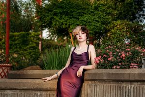 A woman in a burgundy silk dress poses on a stone wall in a garden with pink flowers and greenery.