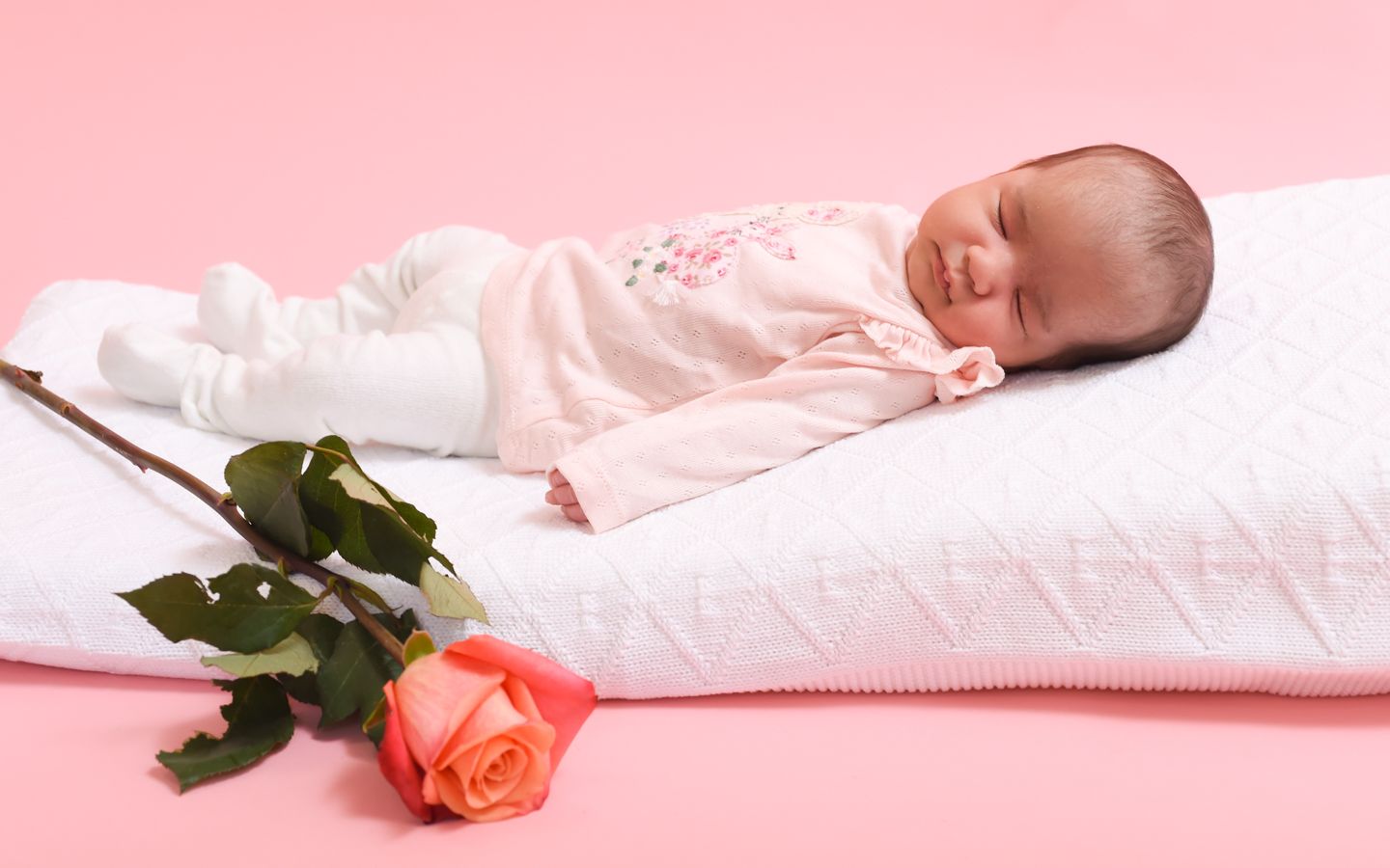 A peaceful baby sleeps on a white blanket, accompanied by a rose, in a newborn photo by Abigail's Studio in Buckinghamshire.