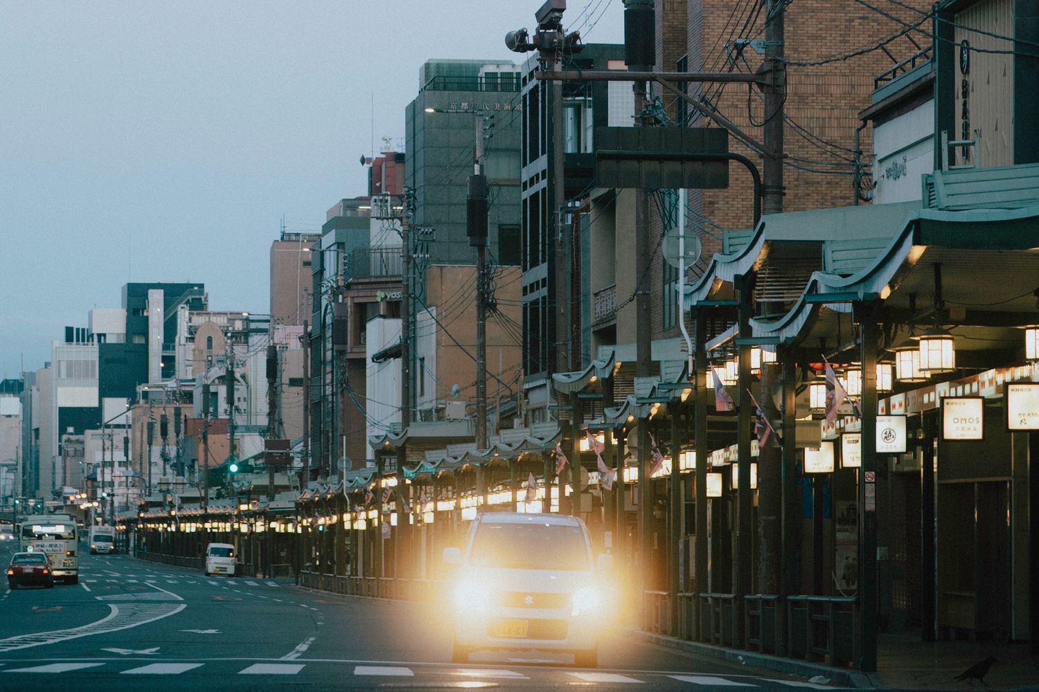 A taxi drives down a quiet Japanese street at dusk lined with traditional lantern-lit storefronts.