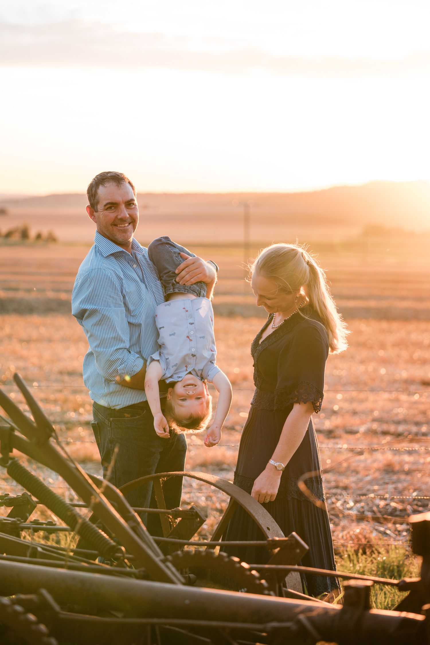 A dad holding his little boy upside down while mum looks on, at sunset on a farm