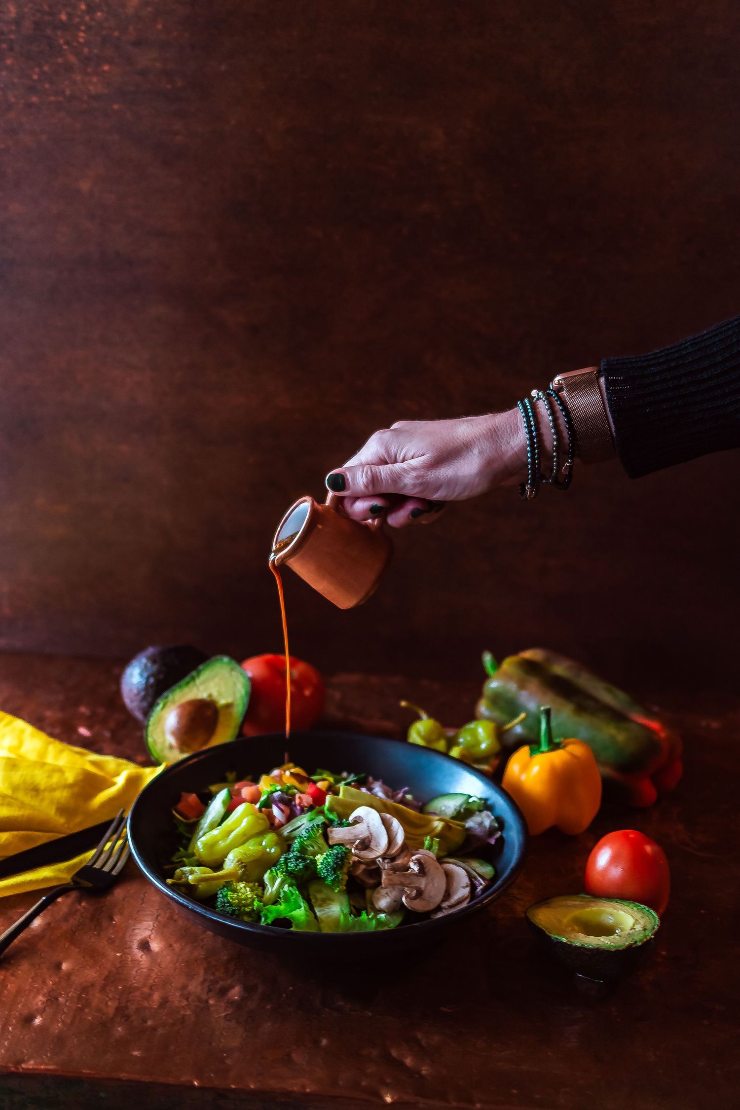 Hand pouring sauce over a colorful salad bowl with mushrooms, avocado, and fresh vegetables on a rustic wooden table.