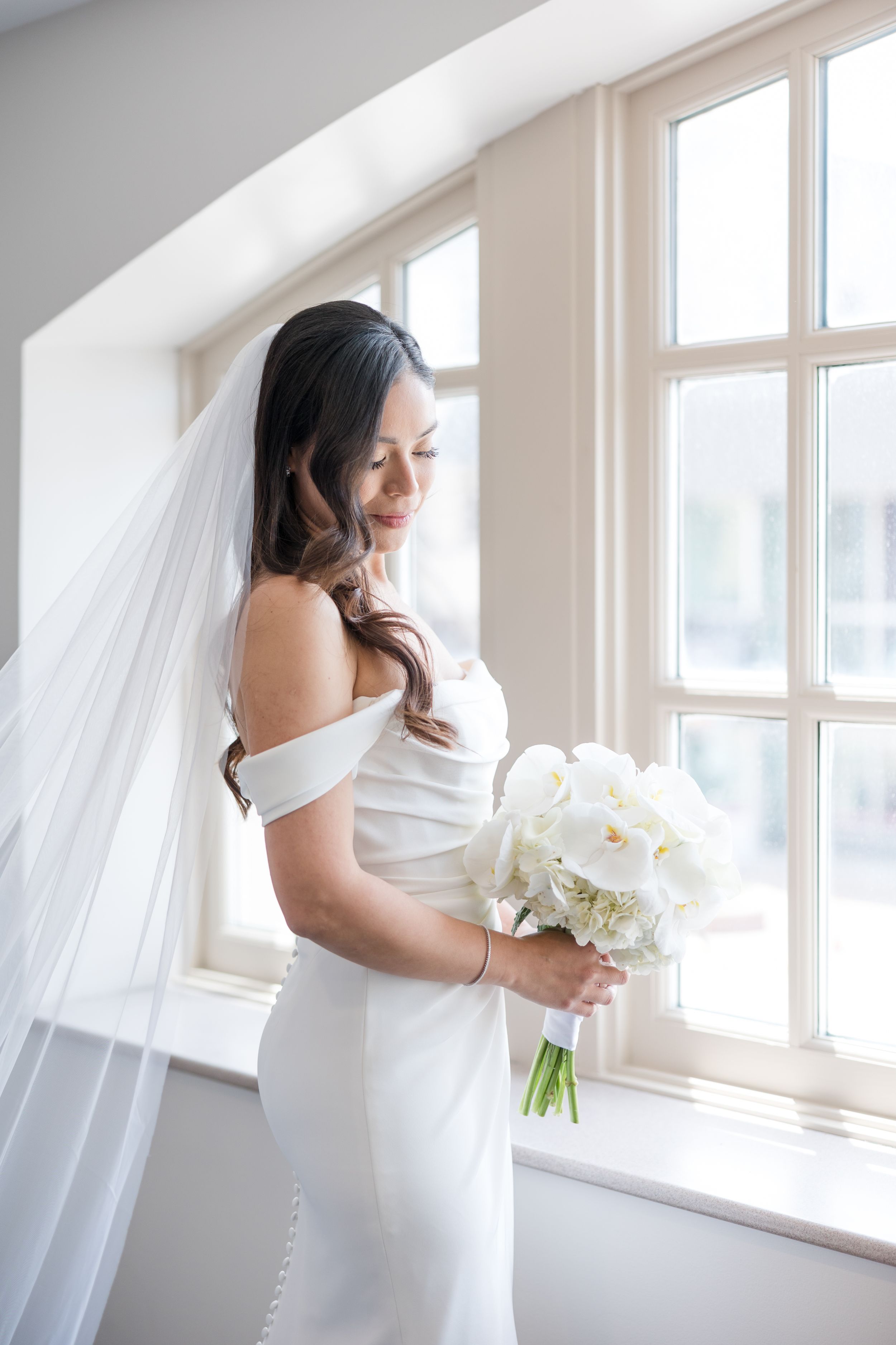 Elegant bride in white off-shoulder dress holds white bouquet by sunlit window.
