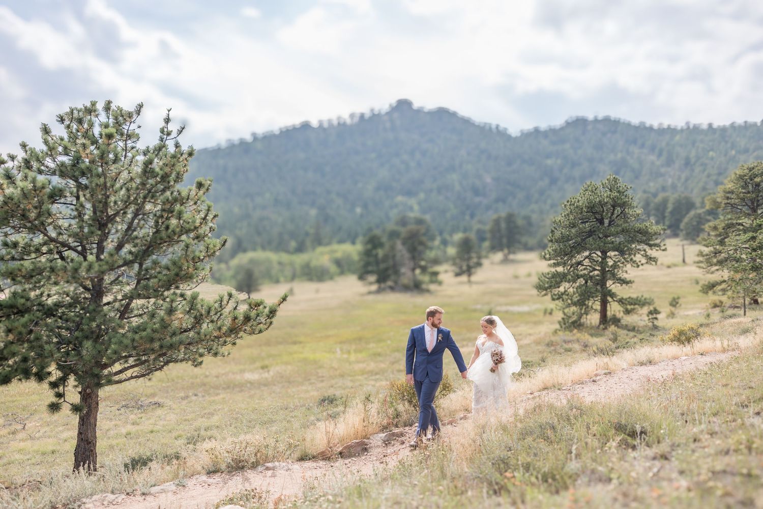 Wedding couple walks along a mountain trail surrounded by evergreen trees and rolling hills in the distance.