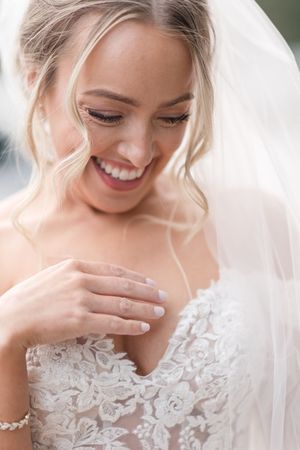 A bride in a delicate lace wedding dress smiles joyfully while wearing a white veil.