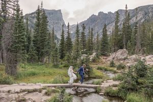 A scenic mountain trail winds through evergreen forest with rocky peaks and cliffs in the background.