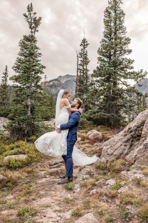 A couple in wedding attire embrace on a rocky mountain overlook surrounded by evergreen trees in the wilderness.