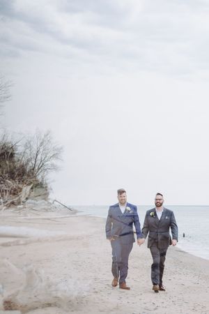 Two people in suits walk hand in hand along a serene beach shoreline on a cloudy day.