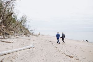 Couple walking along a serene beach with driftwood and sand dunes on an overcast day.