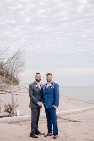 A couple in formal suits stands together on a sandy beach with driftwood and cloudy skies in the background.