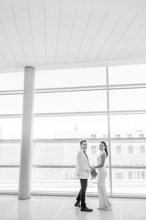 A couple in white attire shares a tender moment in a bright, minimalist space with white walls and columns.