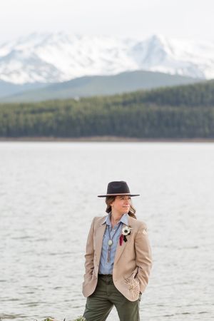 Person in tan jacket and hat poses against mountain lake landscape.