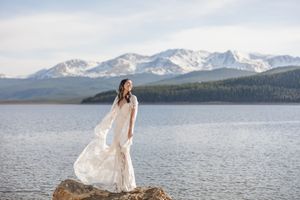 Flowing white dress catches wind against dramatic mountain lake landscape.