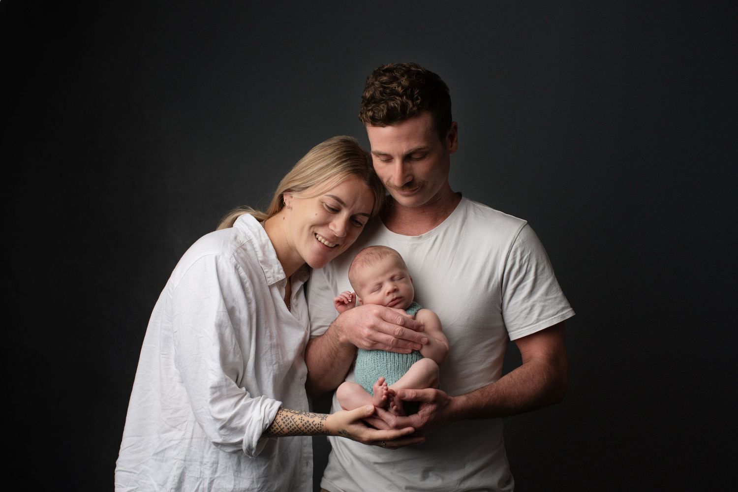 Couple dressed in white holding their newborn baby against a dark studio backdrop in an intimate family portrait — taken in Palmerston North