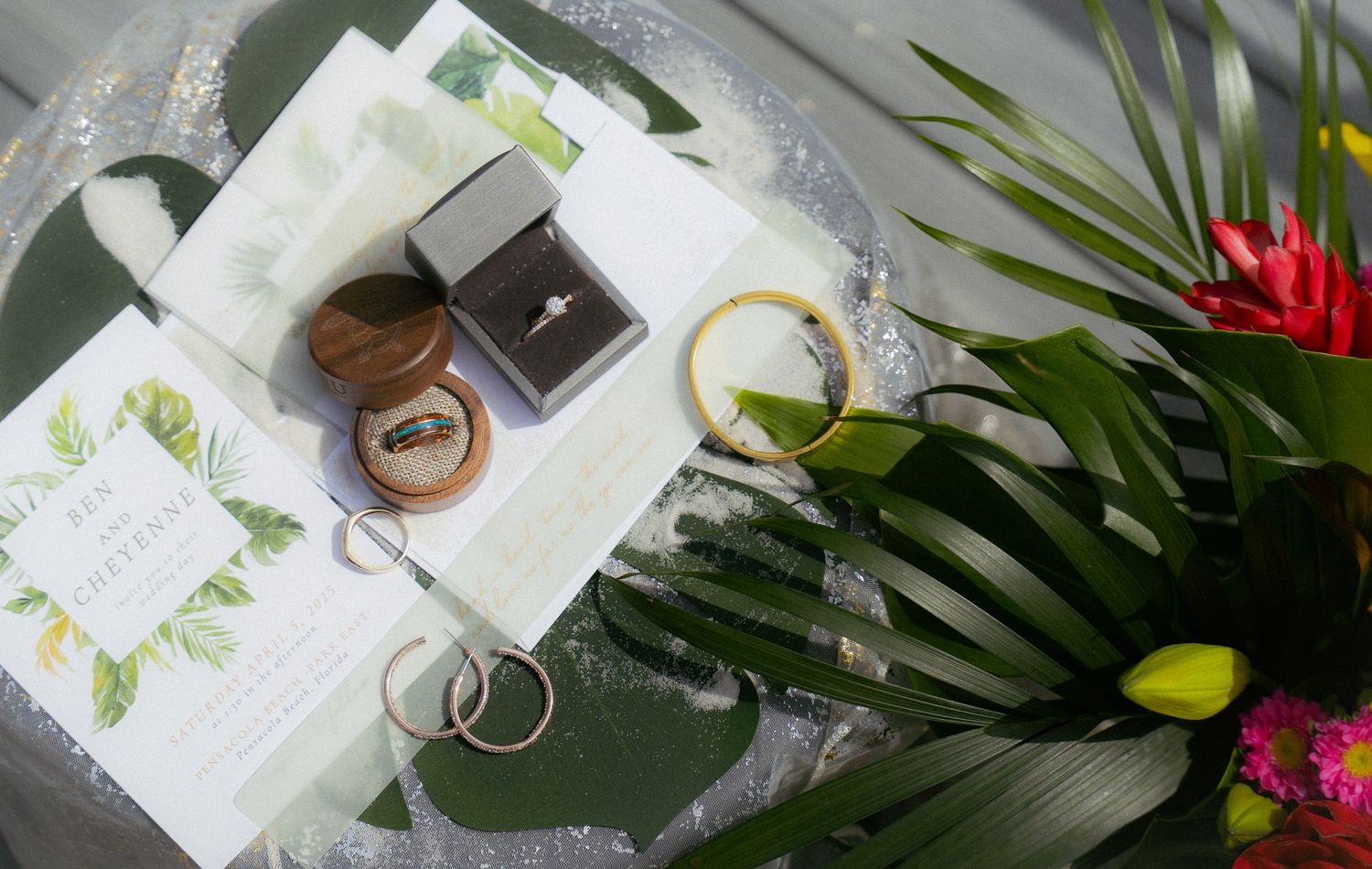 Wedding rings, invitation cards, and decorative elements arranged on a reflective surface with tropical foliage and flowers.