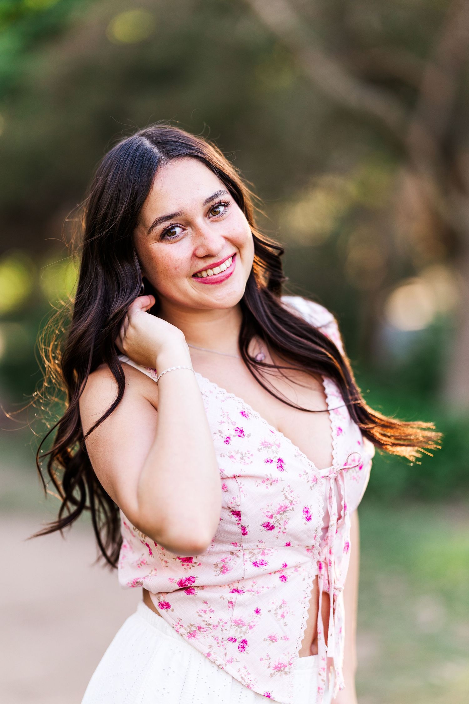 A person in a pink floral top poses outdoors with flowing dark hair and natural lighting in the background.