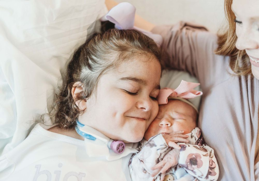 Two children lay close together on white bedding wearing soft pastel colors showing peaceful expressions.