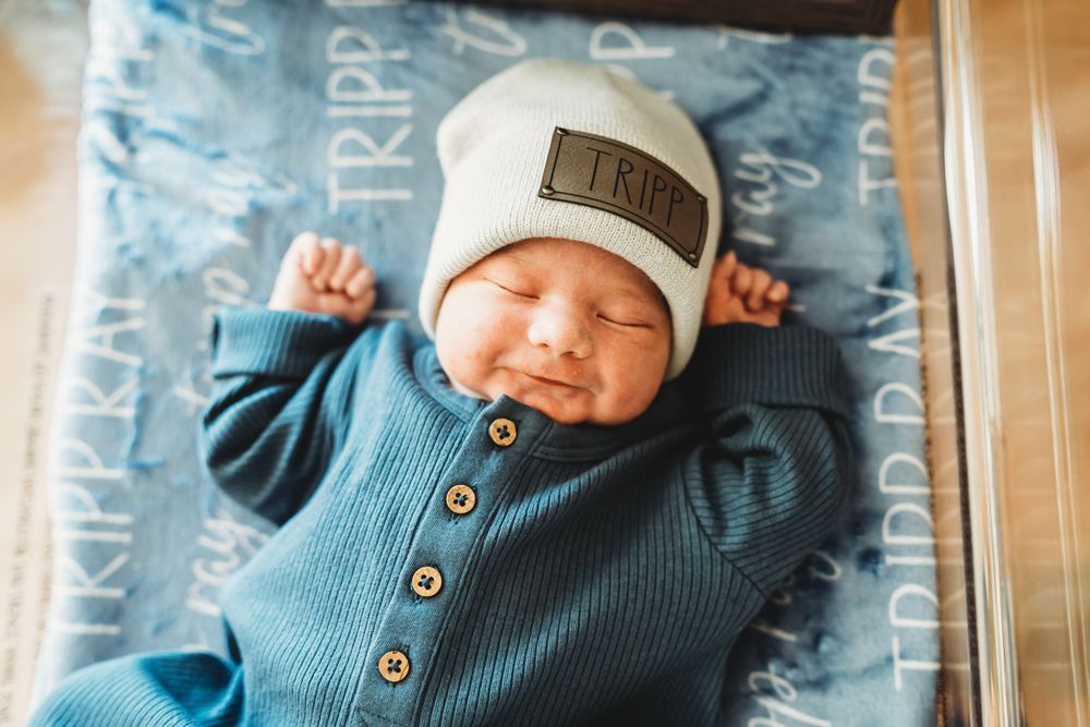 A sleeping newborn in a blue ribbed outfit and white beanie hat stretches while lying on patterned bedding.