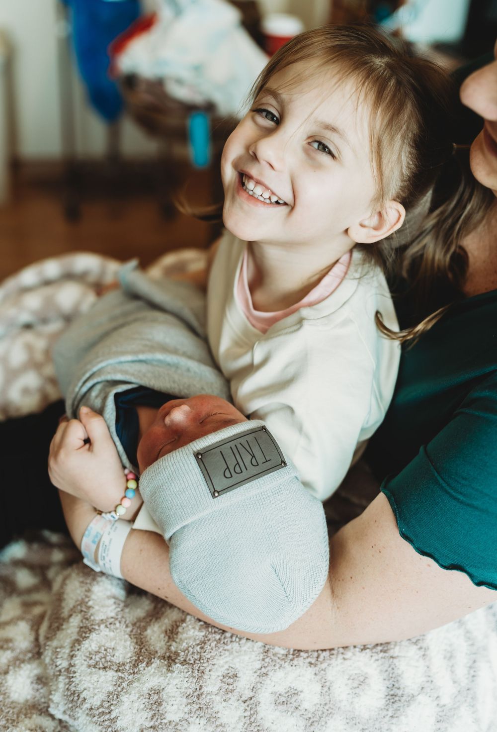 A child wearing a light colored sweater sits on a carpeted floor, smiling brightly while playing indoors.