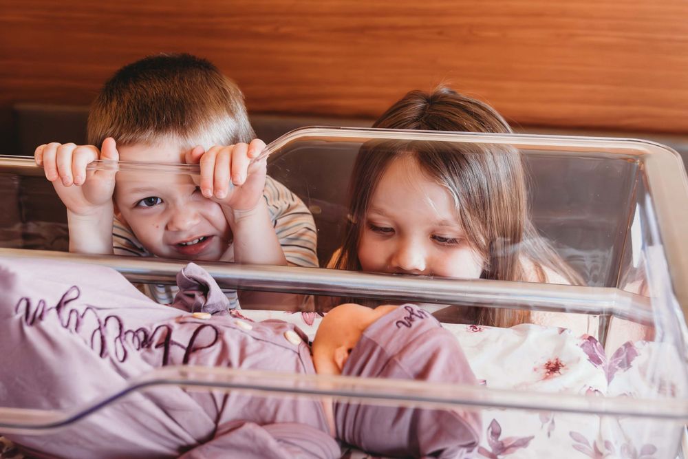 Two children peeking playfully over the edge of a bed with pink bedding.
