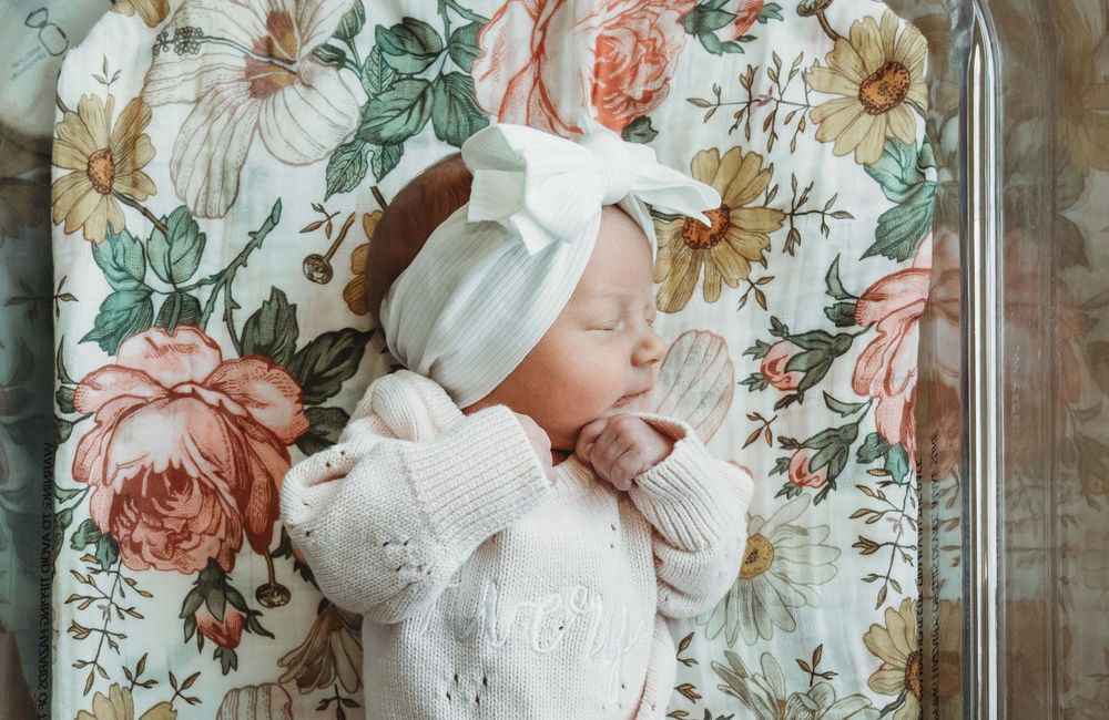 A sleeping baby wearing white clothing and a white bow headband lies on floral fabric with pink and green patterns.