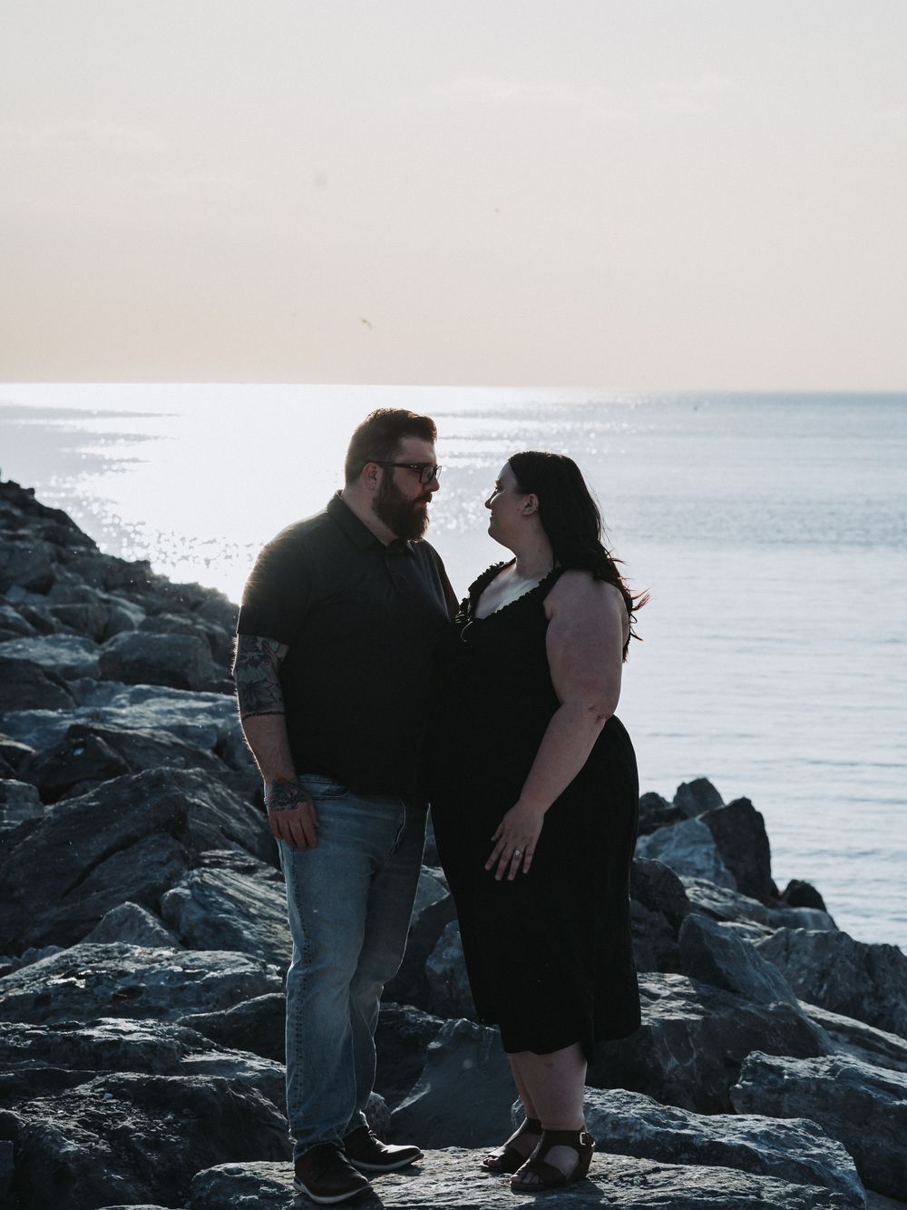 Romantic couple standing on rocky beach at sunset with ocean waves in background.