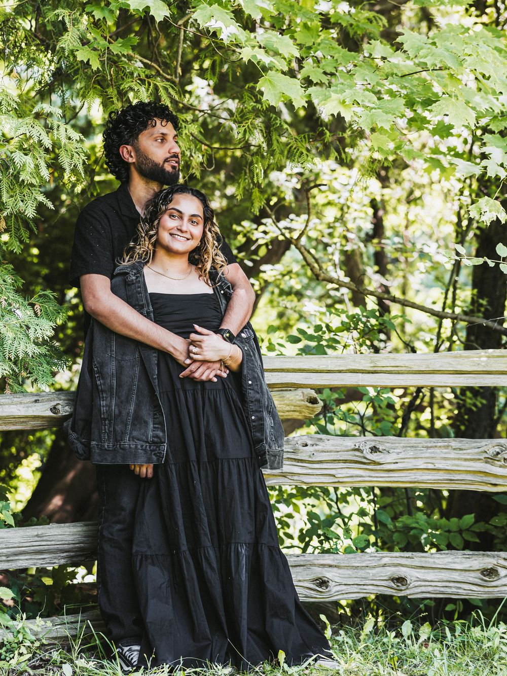 Couple in matching black outfits embrace near a wooden fence surrounded by lush greenery.