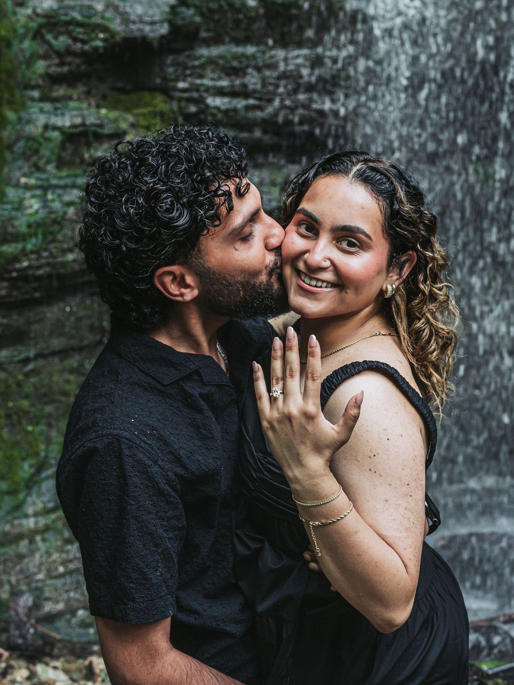 Couple in black clothing showing engagement ring while embracing near waterfall.